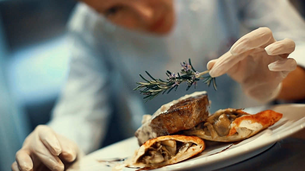 Closeup tilt of blurry female chef placing rosemary on a steak meal before serving. She's using protective gloves when dealing with ready to eat food.